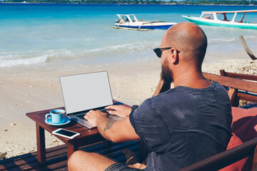 Fototapete - Back view of young man sitting with laptop computer on sea shore working online during vacations, professional male freelancer keyboarding on netbook with mock up screen enjoying summer trip