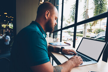 Fototapete - Young bearded guy sitting in cafeteria with laptop computer typing text for publication in blog,skilled male freelancer doing distance job via netbook with mock up screen and wireless internet