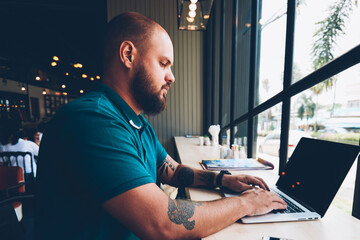Fototapete - Young beared man keyboarding on laptop computer with mock up screen doing remote job sitting in cafeteria, skilled male typing feedback about cafe on netbook working on freelance publicating in blog