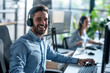 © eshana_blue - Young enthusiastic customer service worker with headset at his desk in a shared office, smiling at camera, his female colleague in the background.