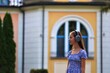 A girl wearing headphones and a blue dress stands in front of a yellow building. The scene is peaceful and calm, with the girl looking away from the camera
