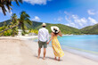 Happy holiday couple with sunhats walks down a tropical beach surrounded by rain forest in the Caribbean, Antigua island