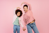 Fototapeta  - Smiling mother and her daughter posing in studio and making heart by their hands, isolated on pink
