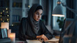© Kafka - Focused Remote Worker: Hijab-Clad Woman Efficiently Managing Tasks on Laptop in Tidy Home Office Setting at Night