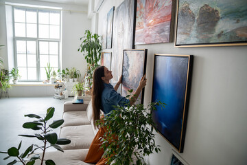 Woman painter hanging newly painted modern framed painting on wall in bright room art workshop with indoor plants. Actual solutions by decoration in contemporary interior design. Artists cosy home