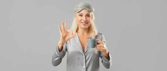 Poster - Mature woman in pajamas with cup of coffee showing OK gesture on grey background