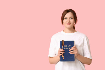 Religious young woman with Holy Bible and rosary beads on pink background