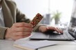 Woman holding tasty granola bar working with laptop at light table in office, closeup