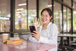 © bongkarn - A cheerful female student sits at a table in a cafeteria talking on a video call with her friend.
