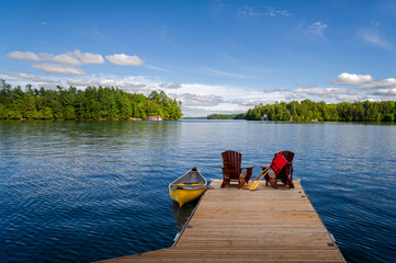 Wall Mural - Two Adirondack chairs on a wooden dock overlook the blue waters of a Muskoka lake in Ontario, Canada. A life jacket and paddles hang from one chair, while a vibrant yellow canoe is tied to the pier.