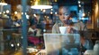 Young African American woman drinks coffee while working with laptop at a cafe. View through the glass of the showcase. Through the glass, a focused young woman commands the nocturnal cafe.