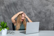 Blond girl in gray dress posing by laptop near fresh green flower