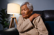senior woman suffering from neck and shoulder pain while sitting on sofa in living room at night