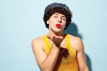 Young happy gay Latin man wear mesh tank top hat clothes with red lipstick make up blow send air kiss isolated on plain light blue background studio portrait. Pride day June month love LGBT concept.