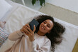 © Westend61 - Joyful young woman lying on bed and smiling at mobile phone