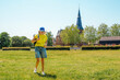 A woman in a yellow shirt and blue shorts walks through a grassy field against the backdrop of an ancient European castle