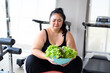 Unhappy overweight Asian female looking for organic green vegetable salad in gym. Asian women in sportswear do not want to eat vegetables. Weight loss workout, healthy lifestyle concept.