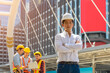 Portrait of engineer or architect in white shirt and safety hat standing in front of the building with group of construction workers on background.