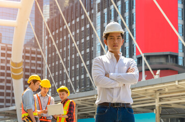 Wall Mural - Portrait of engineer or architect in white shirt and safety hat standing in front of the building with group of construction workers on background.