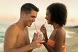 Multiethnic couple sharing a cocktail on the beach - Romantic sunset moments - Joyful interaction by the sea.