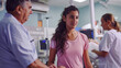 © Natalia - A young woman in a pink shirt talking with an older male doctor in a busy hospital room.