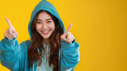 Sticker - Cheerful young Asian woman in blue hoodie pointing towards camera on yellow background