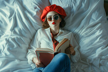 Girl In A Red  French Beret And Sunglasses Lying On A Bed Reading A Book, With Red Lips, Wearing A White Shirt With A Scarf Or Tie And Blue Jeans, In Natural Light