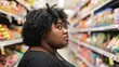 © iuricazac - A woman with curly hair wearing a black top standing in a supermarket aisle looking to her right with a thoughtful expression.