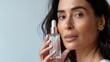Woman with long dark hair holding a clear bottle of skincare product applying it to her face with a gentle touch against a soft-focus background.