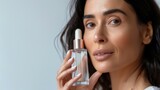 Fototapeta  - Woman with long dark hair holding a clear bottle of skincare product applying it to her face with a gentle touch against a soft-focus background.
