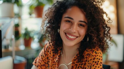 Poster - Smiling woman with curly hair wearing an orange leopard print top sitting in a room with plants and a blurred background.