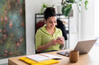 © Studio Marmellata - A content Hispanic woman in a green-striped shirt enjoys her smartphone at a modern workspace, reflecting a seamless blend of technology and lifestyle