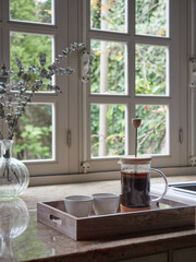 French press coffee maker, with infusion and two cups ready to serve on a tray on the kitchen counter, in front of the window inside the cottage.