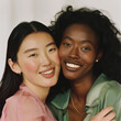 one woman is chinese, the other is black, 2 women hugging and smiling, close crop, one in a pink top and one in a green top, in studio, no makeup and fresh looking