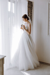 A bride is standing in front of a window, holding a camera and looking out. She is wearing a white dress and a veil