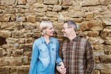 Fototapeta Panele - A happy couple, a 50-year-old man and a 40-year-old woman, stand outside in front of a stone wall, smiling and gazing into each other's eyes