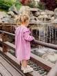 Child girl in stylish dress walking in summer park and looking at waterfall