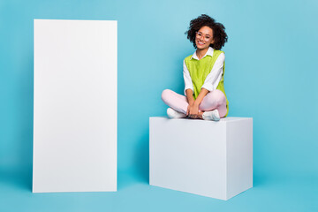 Poster - Photo of charming brown haired lady sit cube podium next to advertisement placard isolated on blue color background