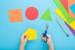 Little child hands holding scissors and cutting colorful geometric shapes from application paper on pastel blue table background. Making different forms. Point of view shot. Closeup. Top down view.