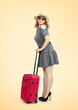 A happy woman in a straw hat, sunglasses, a gray dress and high heels, stands with a red suitcase on a yellow background