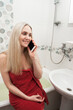 © Ihor - A beautiful smiling blonde girl in a red towel sits in the bathroom, talking on the phone and laughing. Image on the theme of everyday life and social contacts.