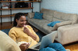 Cheerful young black woman with dreadlocks watching comedy movie on laptop at home, sitting on chair by coffee table, eating popcorn and laughing. African american lady having fun during isolation