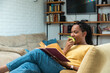 Simple living. African American woman reading book and eating apple in living room. Healthy life and enjoyment at home in free time. Book lover quality time