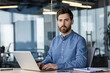 © Tetiana - Portrait of a serious young man working in the office at the laptop, sitting at the desk and confidently looking at the camera