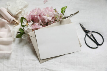 Wedding spring stationery. Feminine scene mockup. Pink blossoming Japanese cherry, apple tree branch. Horizontal blank paper greeting card, black scissors. White linen table background. Flat lay, top