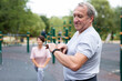 Ssenior man in sportswear looks at fitness watch or at pedometer while exercising in city park