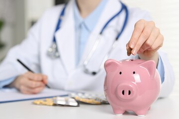 Poster - Doctor putting coin into piggy bank at white table indoors, closeup