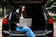 Young woman sitting in car trunk with laptop in nature.