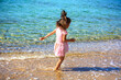 Happy little girl running along the beach back to the camera in summer. Baby in a pink dress