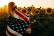 Beautiful young woman with the American flag in a sunflower field at sunset. 4th of July. Independence Day.	